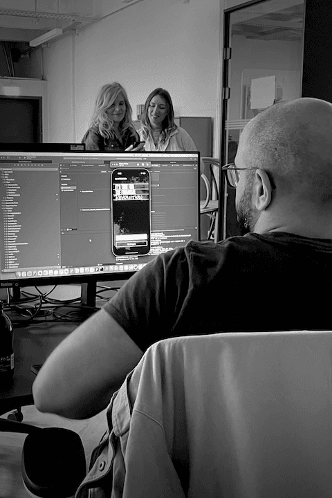 Mann sitzt vor einem Monitor, im Hintergrund stehen zwei Frauen