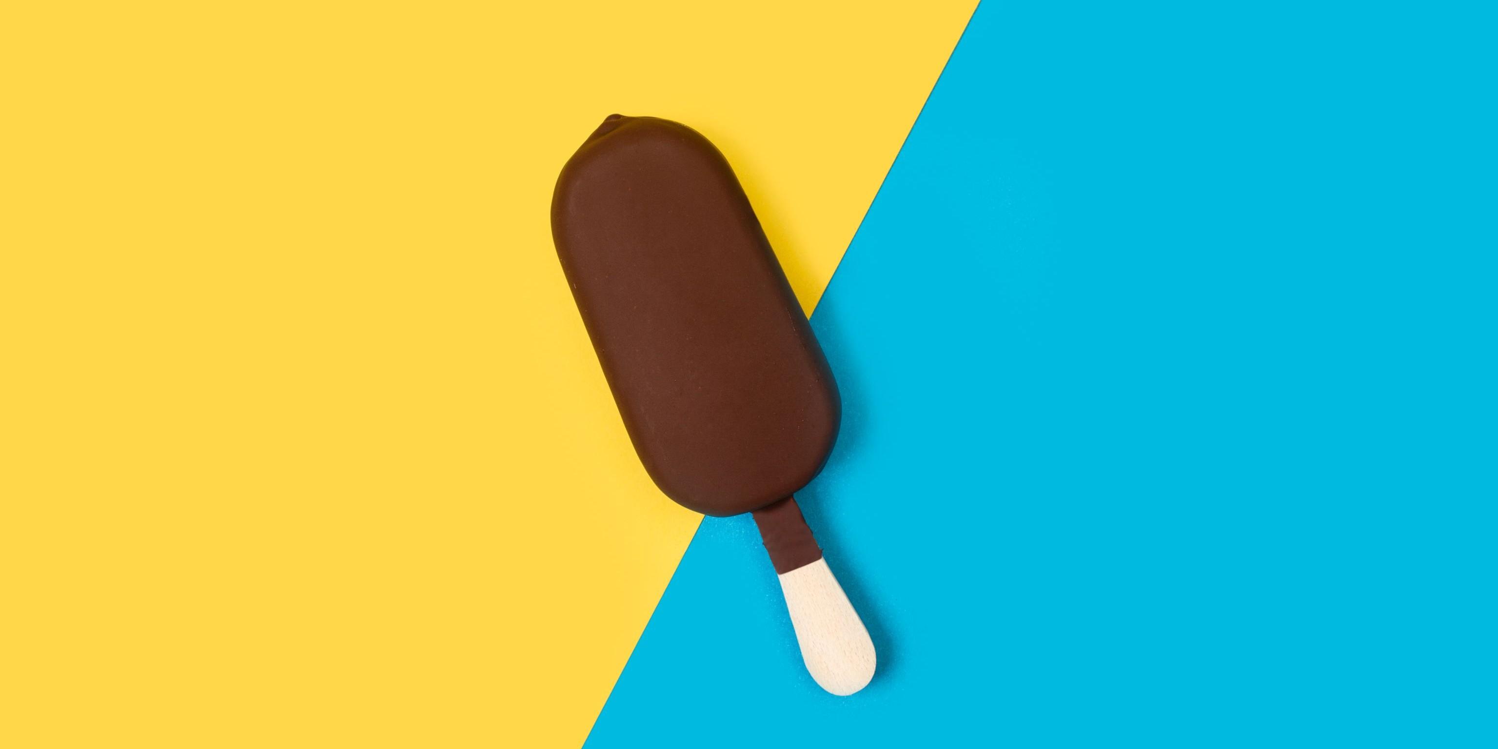 Ein mit Schokolade überzogenes Stieleis vor gelb-blauem Hintergrund